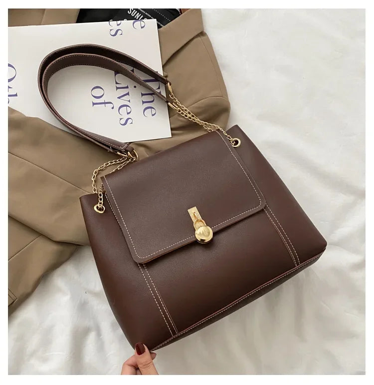 Brown leather handbag with gold chain strap and front clasp, styled on a white surface.
