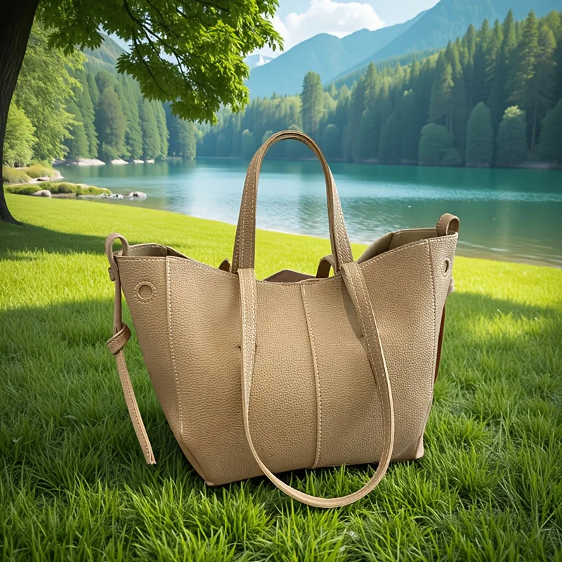 Tan leather tote bag with dual handles resting on grass near a lake.