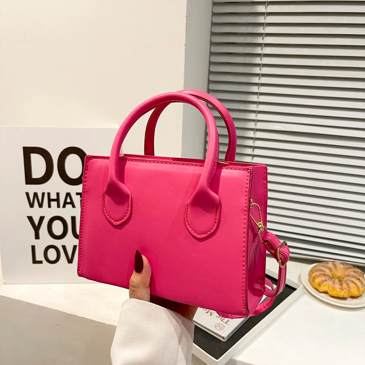 Pink handbag with top handles and a detachable strap, held in front of a decorative sign.