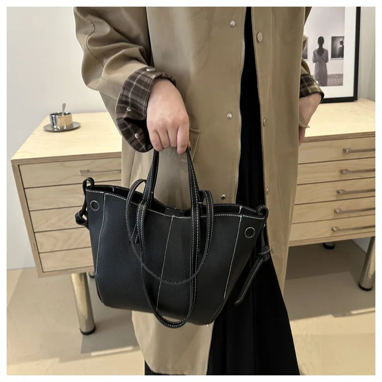 Black tote bag with white stitching and dual handles, held by a person in a beige coat.