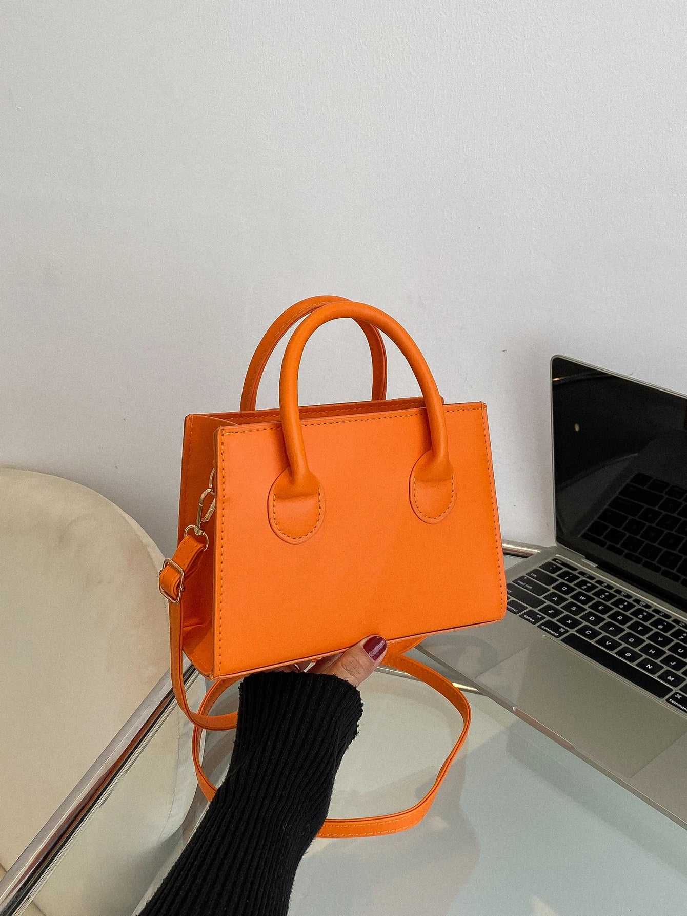 Orange handbag with top handles and a detachable shoulder strap, held against a laptop background.