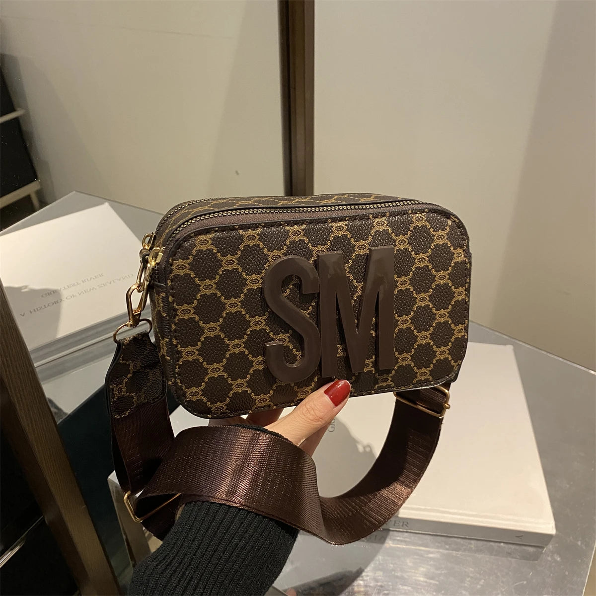 Brown patterned handbag with SM logo and wide strap, held in hand.